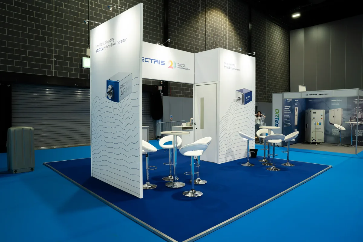 Empty exhibition booth with white partition walls and blue carpet featuring high stools and round tables, showcasing technology displays and branding for a 4D STEM hybrid-pixel detector.