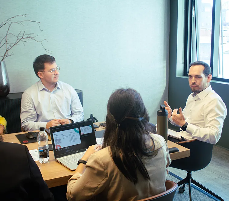 Three professionals in a meeting room, one presenting information on a laptop screen while others listen actively.