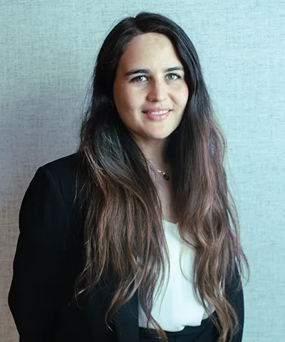 Woman with long dark hair wearing a black blazer and white top standing against a light gray textured background.