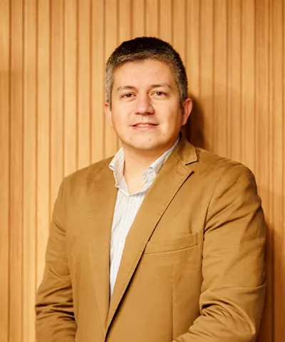 Man wearing a tan blazer and white shirt standing against a vertical wooden slat wall background.