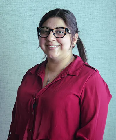 Smiling woman with dark hair in a ponytail wearing large black glasses and a red blouse against a gray textured background.