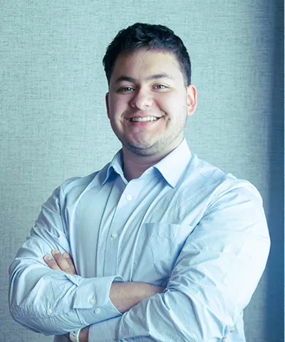 Smiling young man in a light blue button-up shirt standing with arms crossed against a textured gray background.