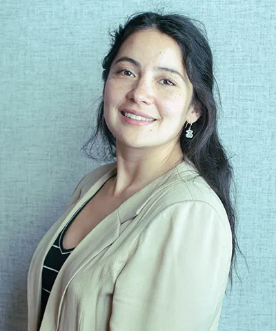 Smiling woman with long dark hair wearing a beige blazer and black top against a textured light blue background.