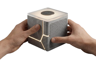 Hands holding a small gray cube-shaped modern speaker with illuminated edges and a circular control on top.