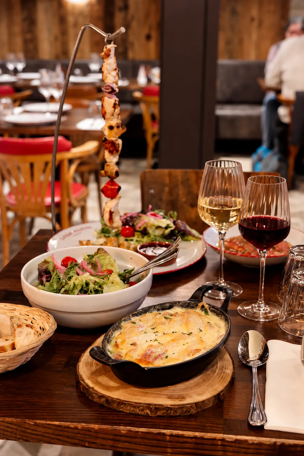 Pique de viande accrochée à un support, avec gratin, salade et verres de vin