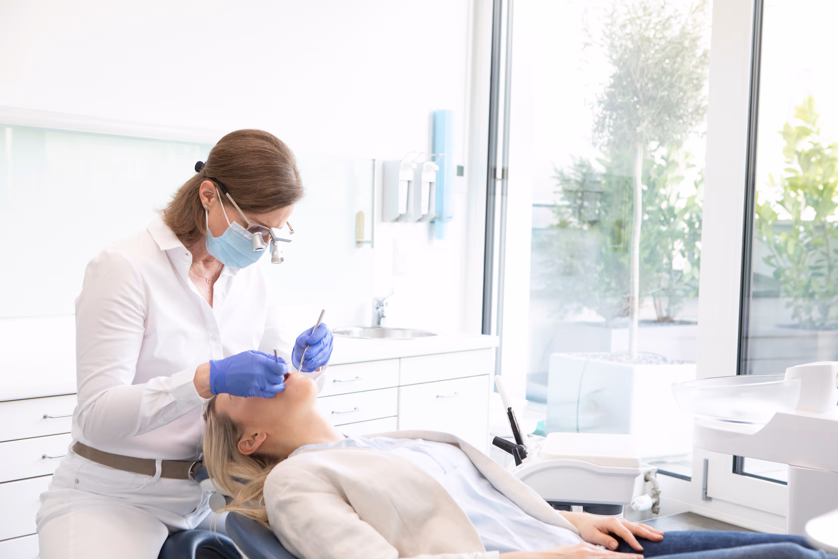 Zahnärztin mit Maske und Lupenbrille untersucht Patientin in heller Zahnarztpraxis.