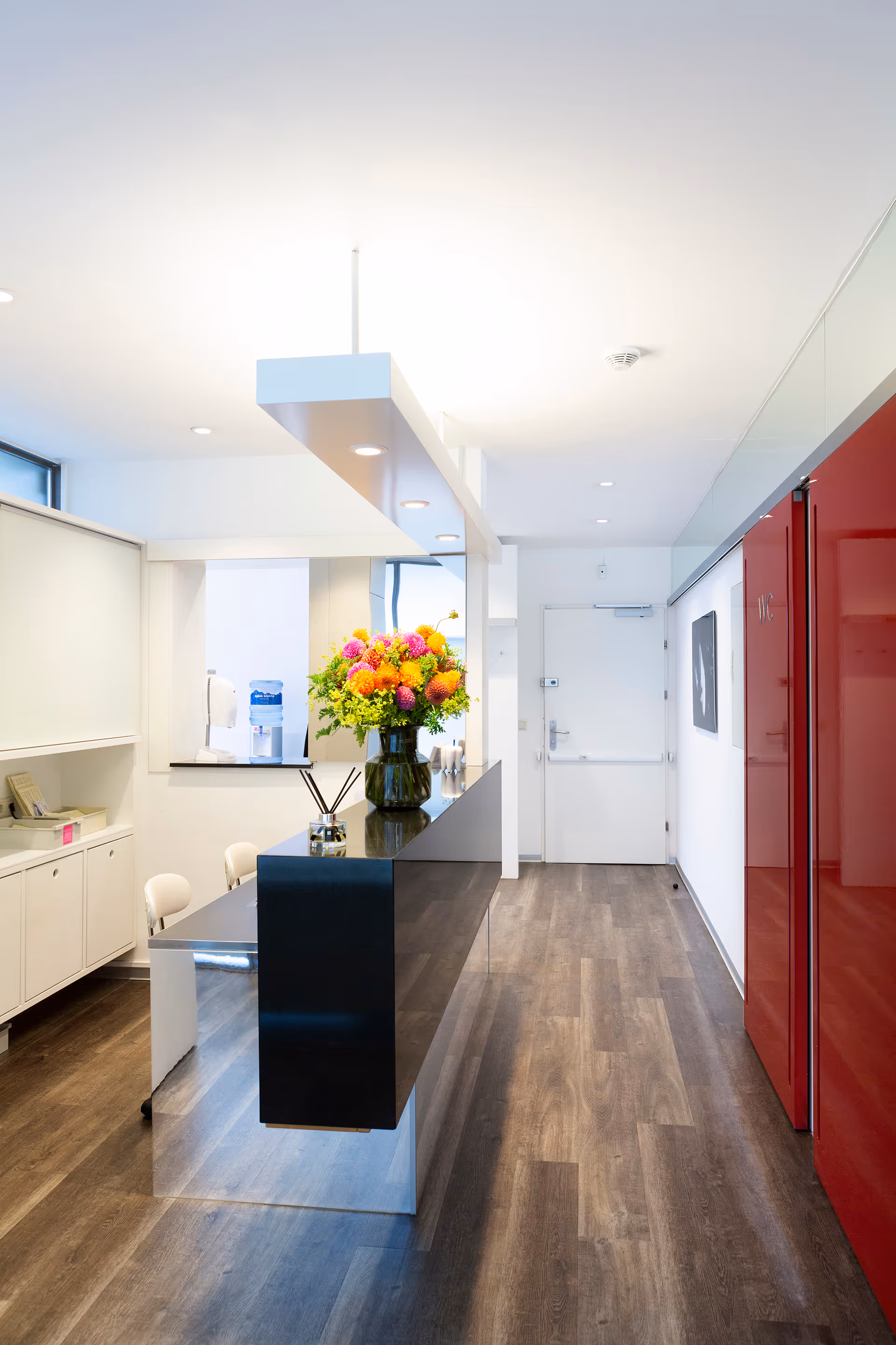 Moderne Empfangstheke mit Blumenvase und roten WC-Türen in einem hellen Büroflur mit Holzboden.