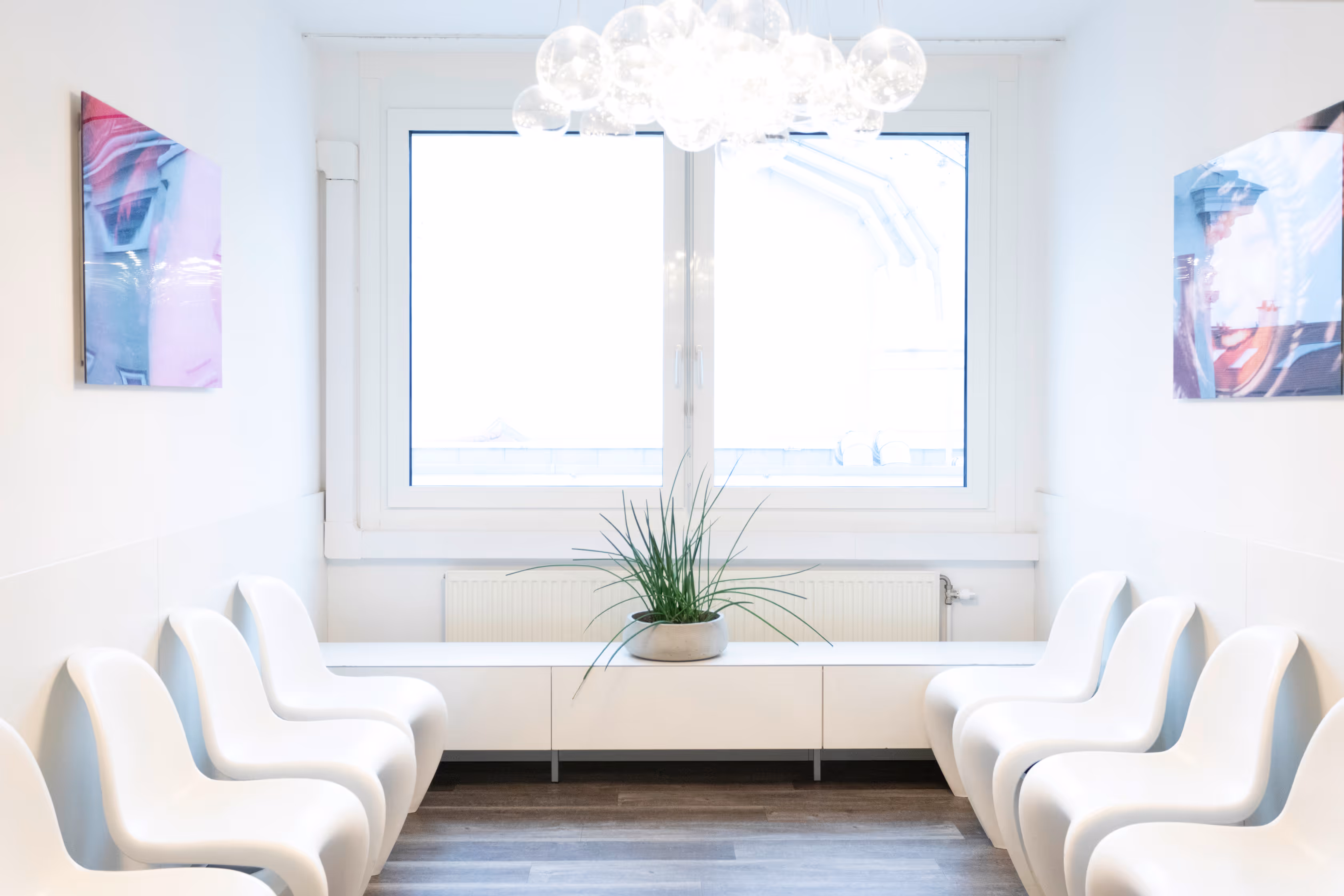 Heller Wartebereich mit symmetrisch angeordneten weißen Stühlen, einem Fenster, einer Pflanze und moderner Deckenbeleuchtung.