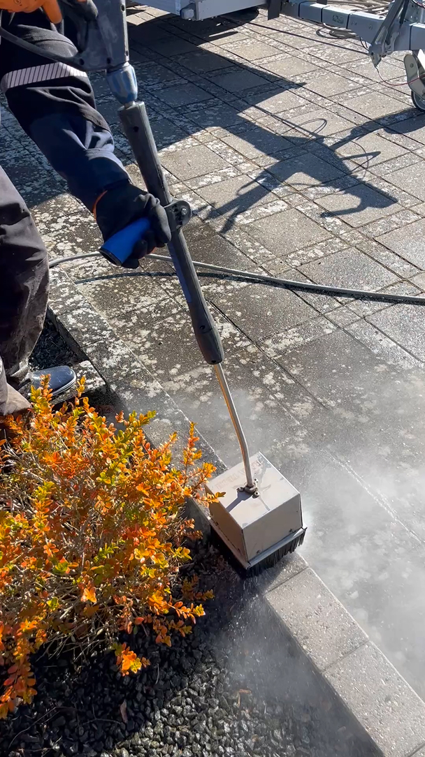 Person rengør fliser med en højtryksspuler nær en busk med orange blade.