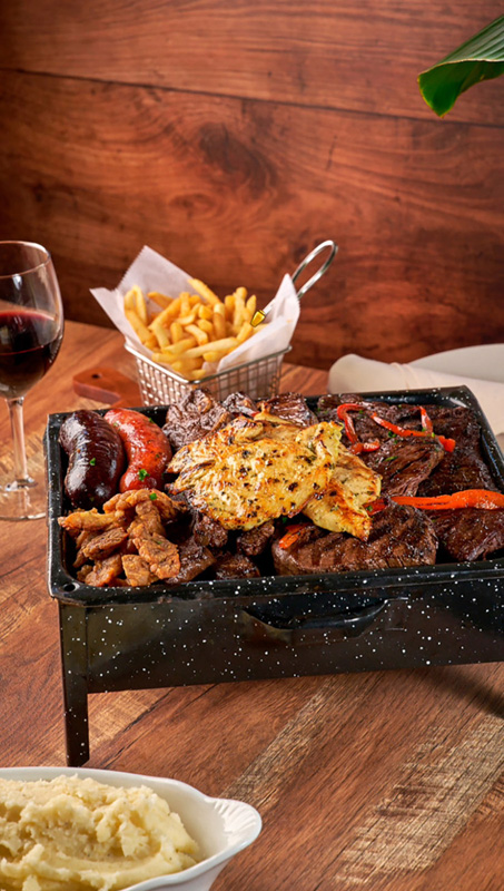 Grilled mixed meat platter with fries in a metal basket, mashed potatoes, and a glass of red wine on a wooden table.