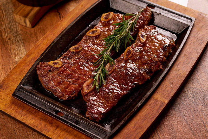 Grilled beef short ribs garnished with a sprig of rosemary on a black serving tray over a wooden board.