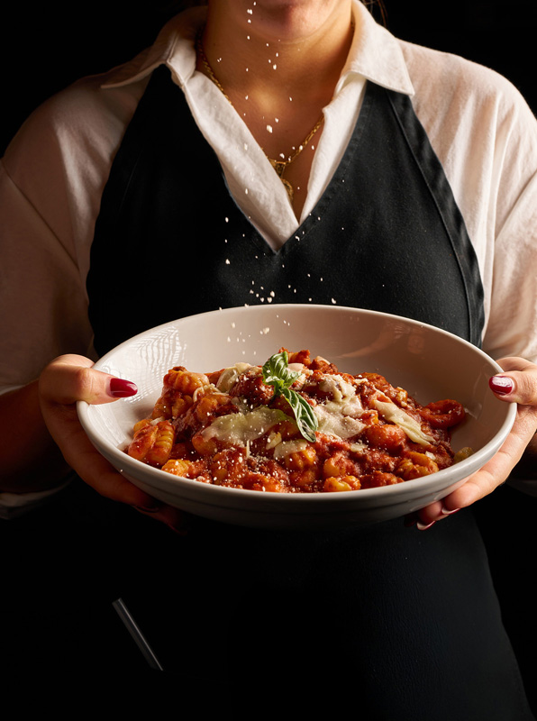 Person in black apron sprinkling grated cheese over a bowl of noqui pasta with tomato sauce and basil garnish.