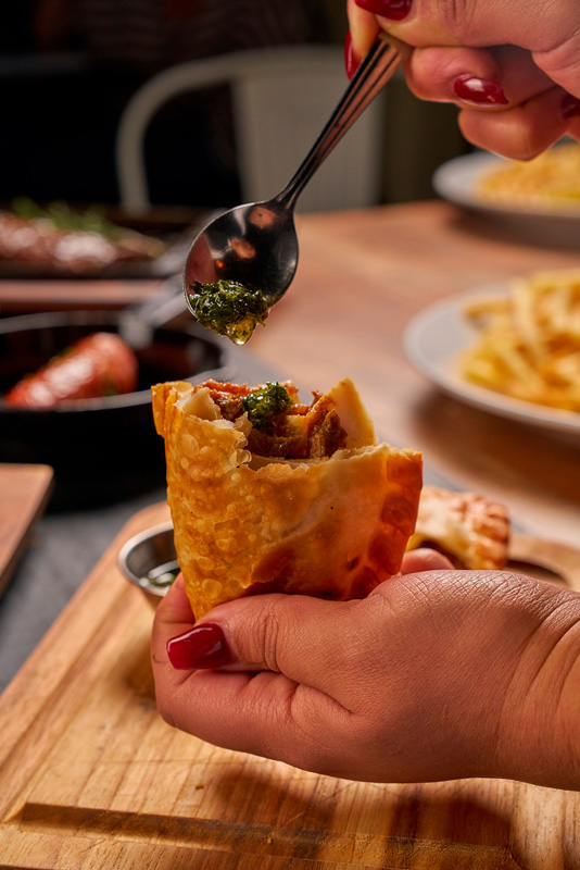 A person holding a partially eaten empanada while spooning green sauce on it, with food dishes blurred in the background.