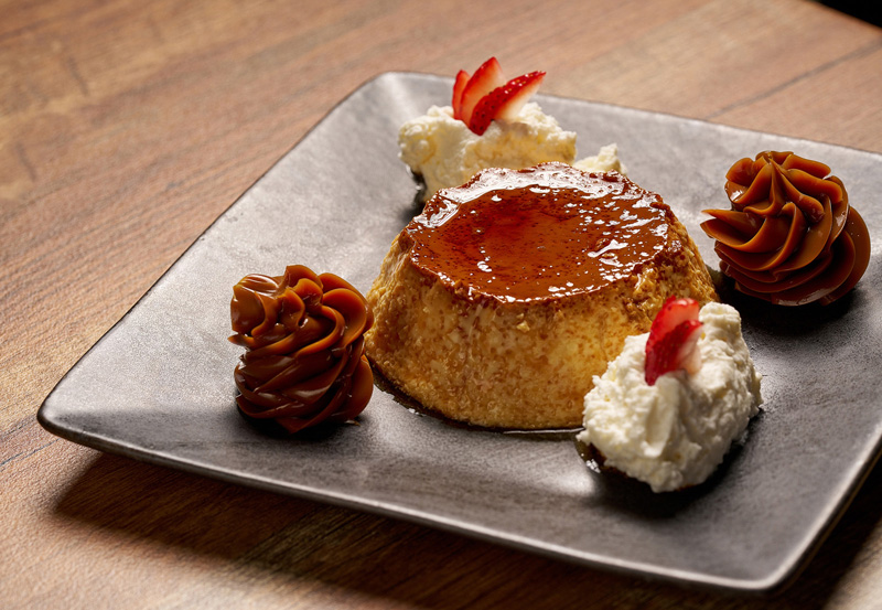 Caramel flan dessert on a square plate garnished with whipped cream and strawberries, alongside two dollops of dulce de leche.