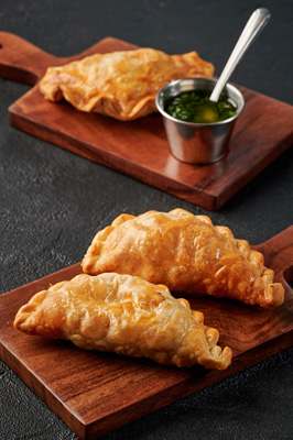 Three golden-brown fried empanadas on wooden boards with a small metal cup of green dipping sauce and a spoon.