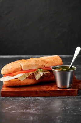 Sandwich with lettuce and tomato on a wooden board next to a metal cup of green sauce with a spoon.