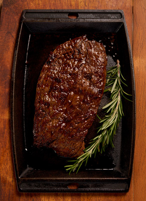 Cooked steak with a sprig of rosemary on a black cast iron tray on a wooden surface.
