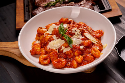 White bowl of gnocchi pasta in tomato sauce, garnished with basil leaves and shaved cheese, on a wooden board.