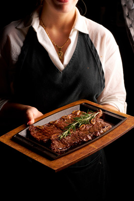 Server holding a wooden tray with a sizzling grilled steak garnished with rosemary.