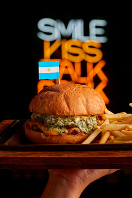 Hand holding a cheeseburger with an Argentine flag toothpick and fries on a wooden tray with blurred neon text in the background.