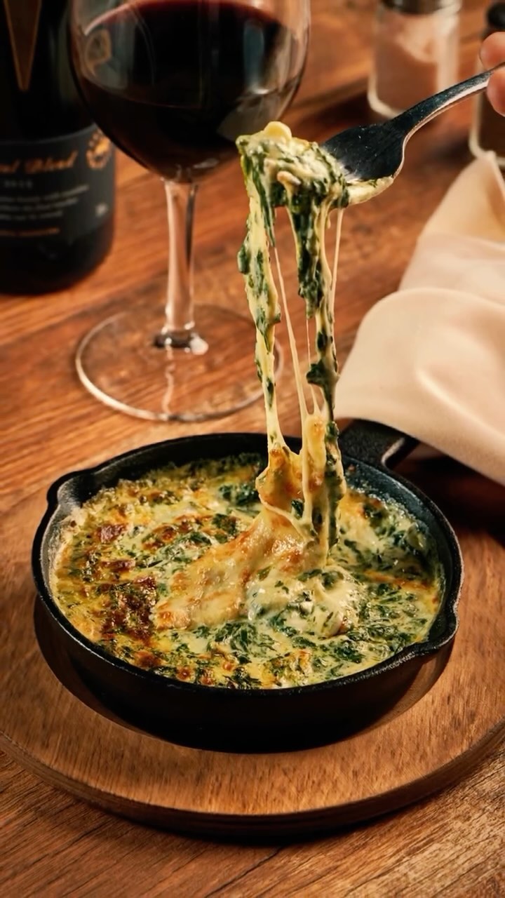Fork lifting melted cheesy spinach dip from a cast iron skillet, with a glass of red wine in the background.