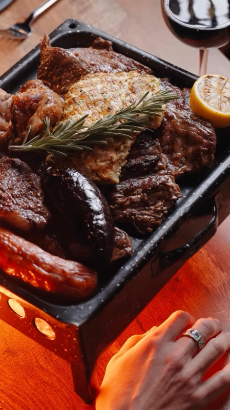 Close-up of a parrillada argentina tray filled with grilled meats including steak, sausage, chicken, garnished with a sprig of rosemary