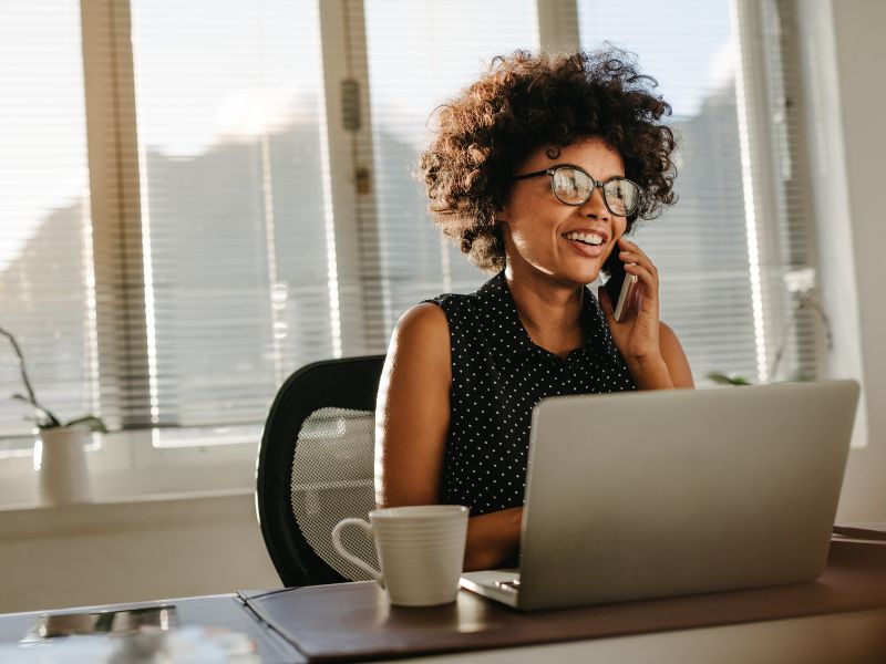 Smiling woman with curly hair and glasses talking on phone while sitting at desk with laptop and coffee cup.