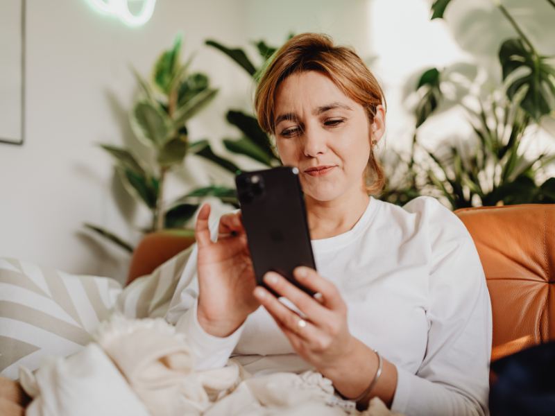 Woman sitting on a couch with plants in the background, looking at her smartphone and smiling slightly.