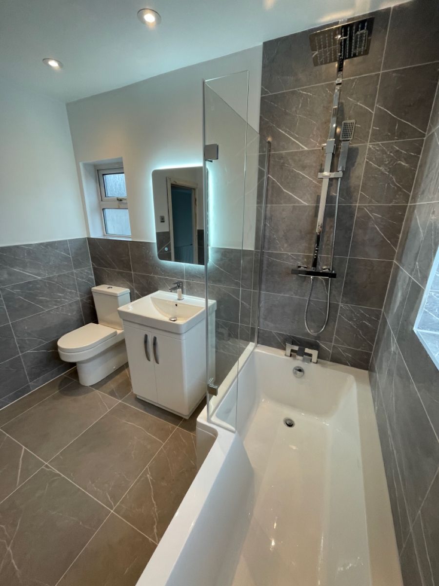 Modern bathroom with white bathtub, shower, sink vanity, toilet, gray tiles, and illuminated mirror.