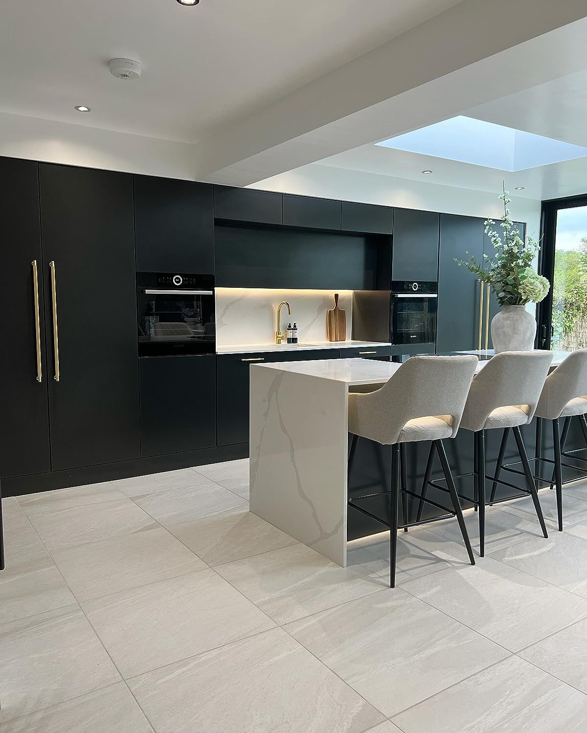 Modern kitchen with black cabinets, white marble island, gray bar stools, and gold fixtures.