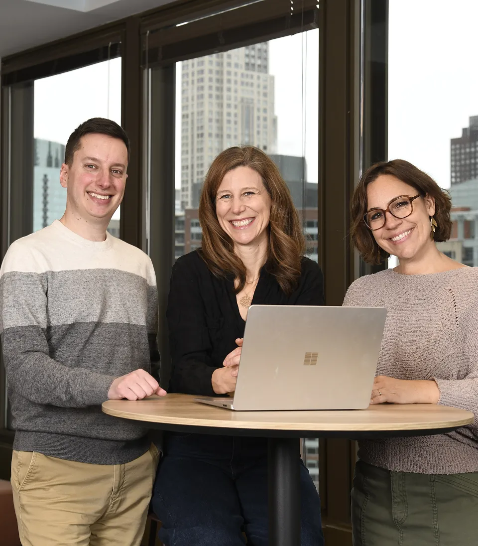Three smiling people standing and sitting around a table with a laptop in front of large windows showing city buildings outside.