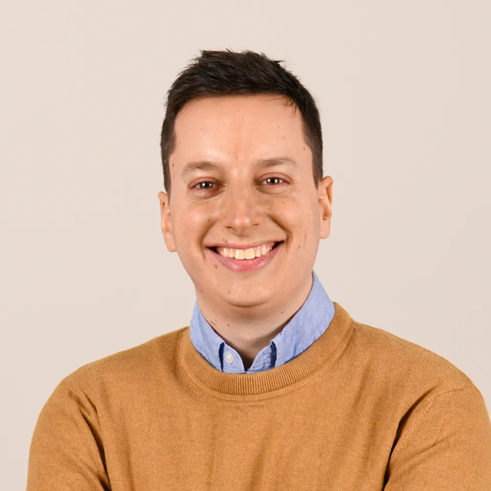 Smiling man with short dark hair wearing a light blue collared shirt and a mustard sweater against a light beige background.