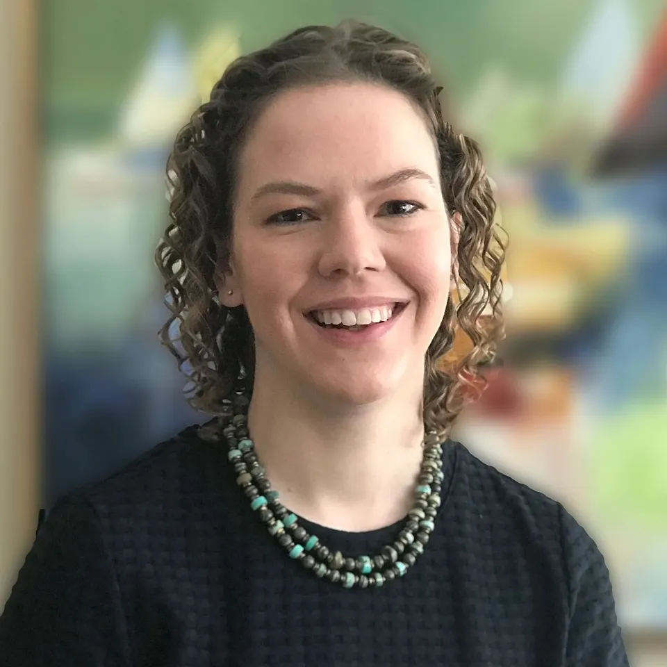 Smiling woman with curly hair wearing a dark top and beaded necklace against a colorful blurred background.