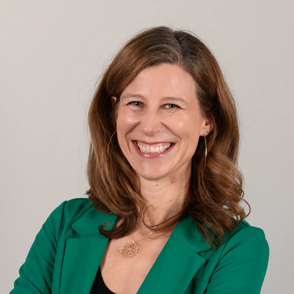 Smiling woman with wavy brown hair wearing a green blazer and a gold necklace against a plain light background.