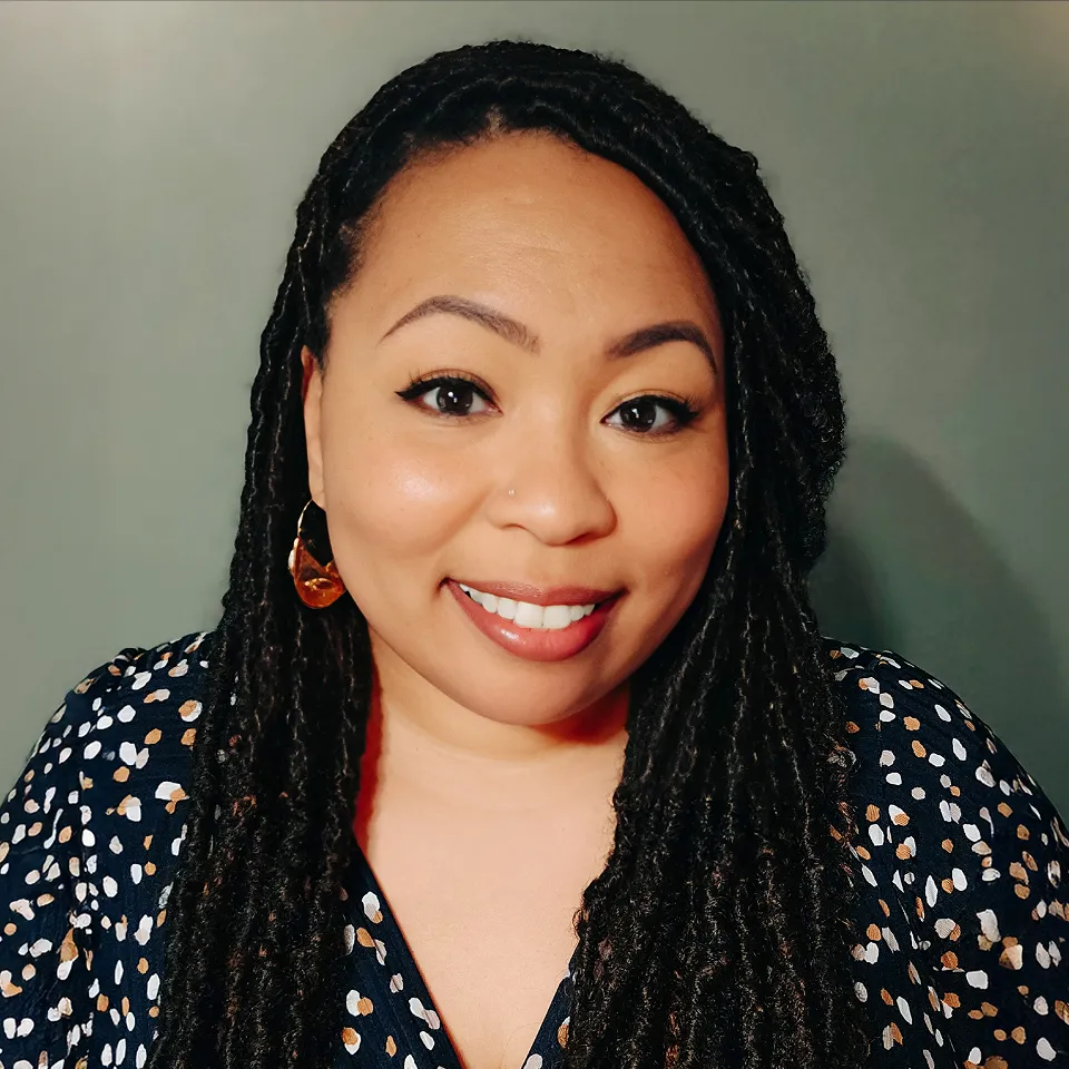 Smiling woman with long braided hair wearing a patterned top and gold hoop earrings against a plain grey background.