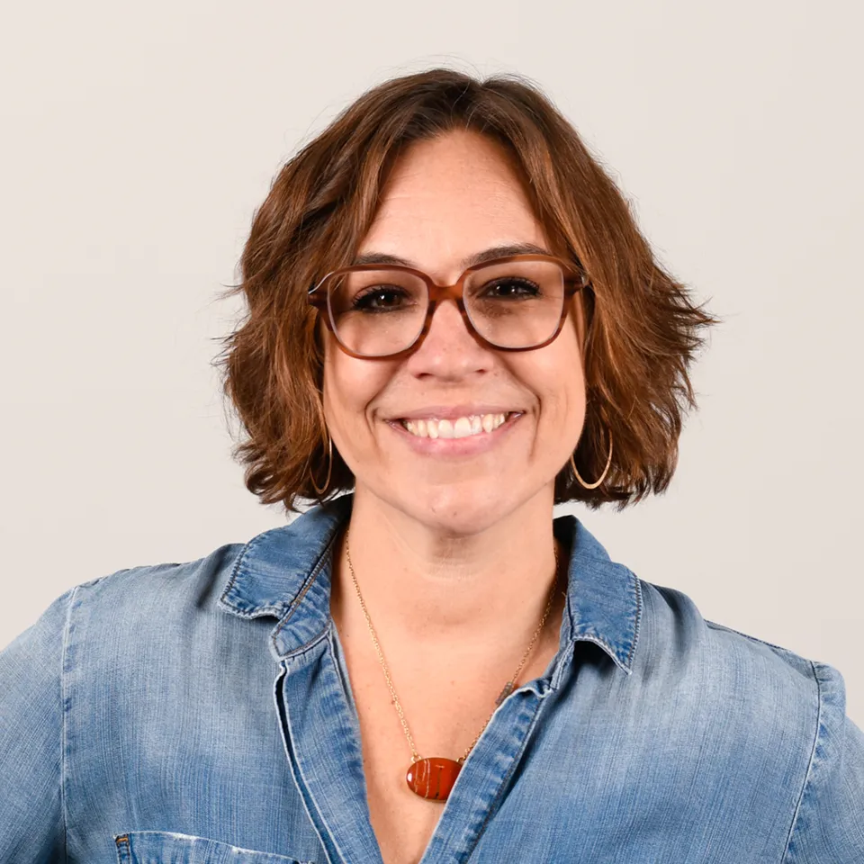 Smiling woman with short brown hair, wearing glasses, hoop earrings, a denim shirt, and a red pendant necklace.