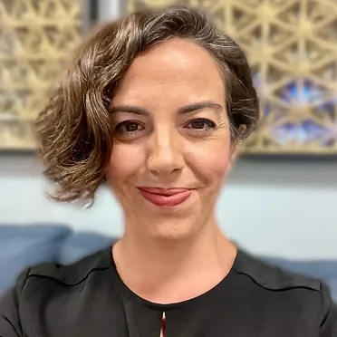Woman with short curly hair smiling in a black top with patterned wall art in the background.