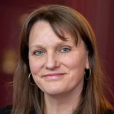 Smiling woman with blue eyes, light makeup, long brown hair, and gold hoop earrings against a blurred maroon background.