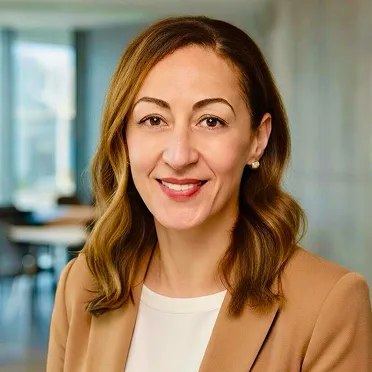 Smiling woman with light brown hair wearing a tan blazer and white top in a modern indoor setting.