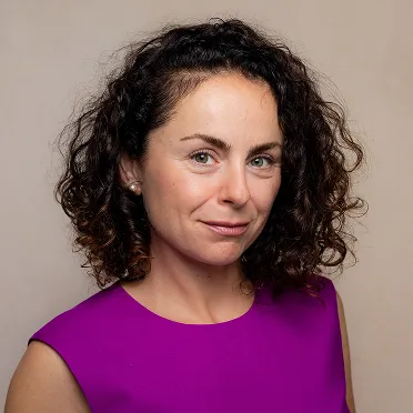 Woman with curly dark hair wearing a sleeveless purple top, smiling slightly against a neutral background.