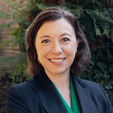 Smiling woman with shoulder-length brown hair wearing a black blazer and green top, standing outdoors with greenery in the background.