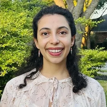 Smiling woman with curly dark hair standing outdoors with green trees and sunlight in the background.