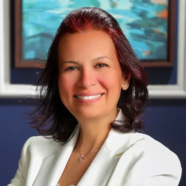 Smiling woman with dark red hair wearing a white blazer and a necklace, standing in front of a colorful abstract painting.