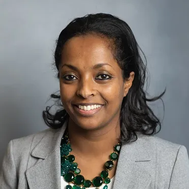 Smiling woman with dark hair wearing a light gray blazer and a green beaded necklace against a gray background.