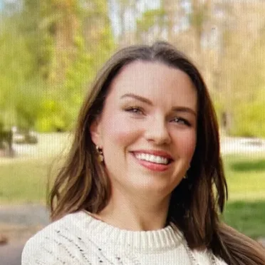 Smiling woman with long brown hair wearing a white sweater outdoors with greenery in the background.