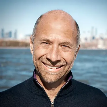 Smiling middle-aged man with a shaved head and beard standing outdoors near water with a cityscape in the background.