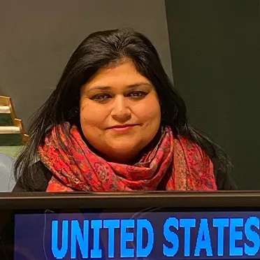 Woman with long dark hair wearing a red patterned scarf seated behind a sign that reads United States.