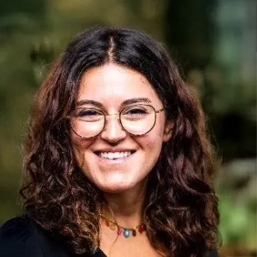 Smiling woman with curly dark hair, round glasses, and a beaded necklace, outdoors with blurred background.