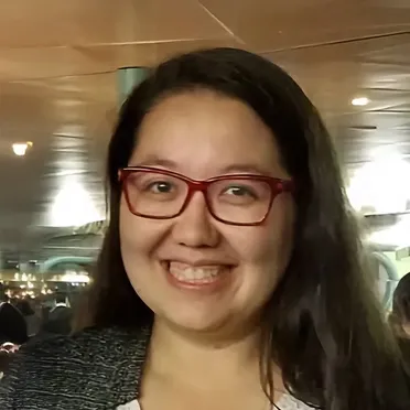 Smiling woman with long dark hair and red glasses in a warmly lit indoor setting.