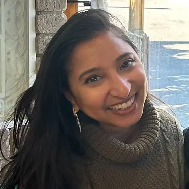Smiling woman with long dark hair wearing a brown knit turtleneck sweater and a dangling earring indoors near a window.